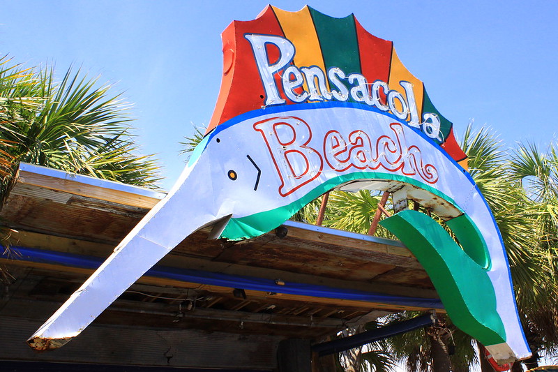 Sign with neon letters spelling out "Pensacola Beach" on swordfish-shaped sign.
